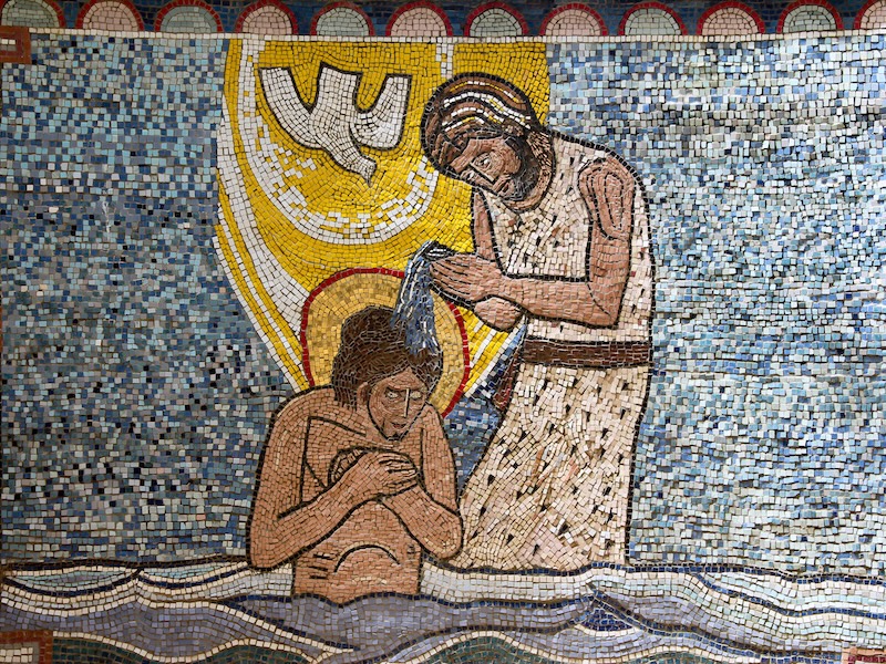 A mosaic of Christ's baptism; standing in a river, John is pouring water over his head; a light shines behind Christ with a dove descending upon him. Blue background.