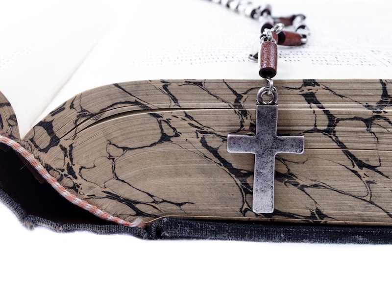 what looks to be a fairly old bible, open and viewed from the edge, with a rosary cross hanging off the side into the edge. plain metal cross, not a crucifix.